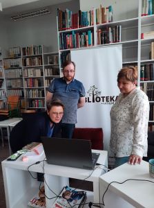 Zdjęcie przedstawia 3 uśmiechnięte osoby - usteckich bibliotekarzy, stojących przed komputerem podczas spotkania online z Tomem Justyniarskim