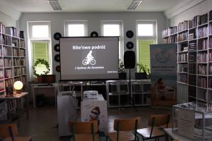 Zdjęcie przedstawia wnętrze Czytelni dla Dorosłych: ekran, regały z książkami i rozstawione krzesła. Na ekranie wyświetlony pierwszy  slajd prezentacji.