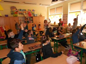 Zdjęcie przedstawia grupę dzieci w klasie szkolnej, które podnoszą ręce w górę podczas spotkania autorskiego on-line z Tomem Justyniarskim